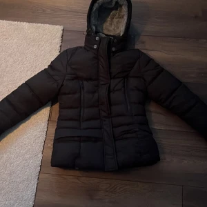 Svart pufferjacka med foder från Marikoo - Säljer en svart pufferjacka från Marikoo med mysigt grått foder och huva. Jackan har långa ärmar och stängs med dragkedja och knappar. Perfekt för riktigt kalla vinterdagar 😊Köpt för 1200kr 