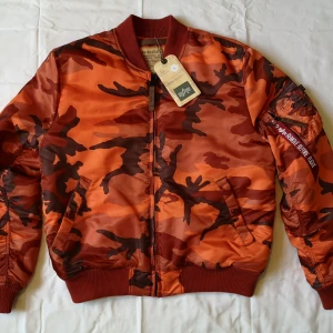 Orange camouflage bomberjacka fr. Alpha Industries  - Säljer en cool orange bomberjacka från Alpha Industries med ett snyggt camouflagemönster. Jackan har dragkedja och ribbade muddar. Perfekt för att sticka ut i mängden! Jackan har använts av endast en person i ett fåtal gånger och kommer från ett djur- och rökfritt hem.