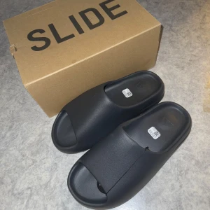 Svarta Yeezy Slide från Adidas - Säljer ett par helt nya Yeezy Slide då dem var för stora på mig. Dessa slides är stilrena och bekväma, perfekta för avslappnade tillfällen. De kommer i originalförpackning.