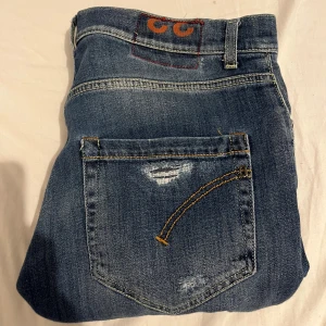 Blå jeans med slitningar - Snygga blå jeans med slitningar och en avslappnad look. De har en klassisk femficksdesign och orange sömmar som ger en extra detalj. Perfekta för en casual stil.