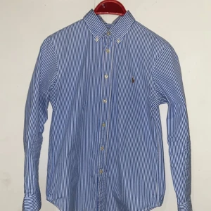 Randig skjorta från Ralph Lauren - Snygg blå- och vitrandig skjorta från Ralph Lauren med klassisk krage och knappar framtill. Skjortan har långa ärmar och en broderad logotyp på bröstet. Perfekt för en stilren look.