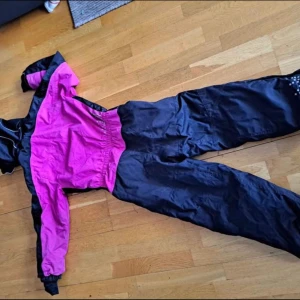 Rosa och svart overall - Säljer en cool rosa och svart overall med dragkedja framtill. Overallen har långa ärmar och är perfekt för kyligare väder. Den har praktiska fickor med dragkedjor och en elastisk midja för bättre passform.