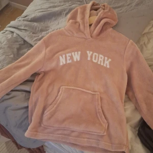 Rosa hoodie med texten 'New York' - Säljer en rosa fluffig hoodie med testen new York. Jätte mysig o lurvig i stl 158/164.  Använd ett fåtal gånger, så inga defekter och ett väldigt bra skick. Säljer pga att den aldrig kommer till användning