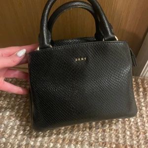 Svart handväska från DKNY - Snygg svart handväska från DKNY med ett elegant ormskinnsmönster. Väskan har två handtag och en praktisk dragkedja upptill. Perfekt för att bära dina viktigaste saker med stil.