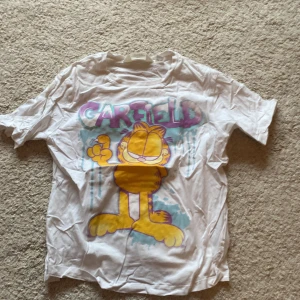 Vit Garfield t-shirt från H&M - Säljer en vit t-shirt från H&M med ett stort Garfield-tryck i färgerna lila, gul och blå. Perfekt för alla Garfield-fans! T-shirten är kortärmad och gjord i bomull.