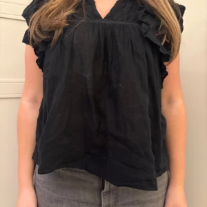 Svart blus med volangdetaljer - Snygg svart blus med volangdetaljer runt ärmarna och halsen. Perfekt för en stilren look. Blusen har en lös passform och korta ärmar.