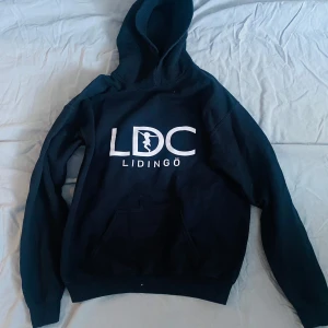 Svart hoodie från Gildan med Lidingö-tryck - Säljer en svart hoodie från Gildan med trycket 'LDC Lidingö' på framsidan. Tröjan har en klassisk design med huva och en stor ficka framtill. Perfekt för en avslappnad stil. Storlek XL, men passar någon med S, då den är gjord i barnstorlek.