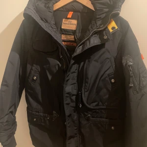 Svart jacka från Parajumpers - Säljer en svart Parajumpers jacka med huva och flera praktiska fickor. Jackan har dragkedja och knappar framtill samt en broderad logga på ärmen. Perfekt för kyliga dagar. Och den här 100% äkta köpt från NK