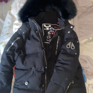 Svart dunjacka från Moose Knuckles - Säljer en svart dunjacka från Moose Knuckles med pälskantad huva. Jackan har långa ärmar och flera fickor. Perfekt för kalla dagar. 1 nackdel är att dragkedjan är lite fuckad men kan visa hur man gör.