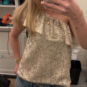 Beige paljettopp med volang - Snygg beige paljettopp med volangdetalj. Perfekt för festliga tillfällen med en glittrig look och offshoulder-design.