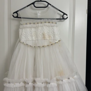 Vit klänning med spets och pärldetaljer - Charmig vit klänning med spetsöverdel och flera lager tyll i kjolen. Klänningen har pärldetaljer runt midjan och längs kjolen samt en stor rosett baktill. Perfekt för speciella tillfällen.behöver tvätt