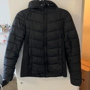 Svart pufferjacka från Etirel - Säljer en svart pufferjacka från Etirel med dragkedja och huva. Jackan är perfekt för kyligare väder och har en stilren design. Tillverkad i nylon och polyester för extra värme och komfort.