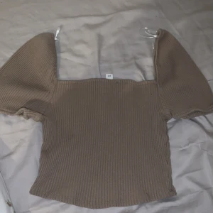 Brun ribbad topp från H&M - Säljer en snygg brun ribbad topp från H&M med puffiga korta ärmar. Perfekt för en stilren look. Toppen har en fyrkantig halsringning och är i ett mjukt material.
