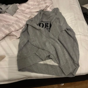 Grå sweatshirt från Morris - Säljer en grå sweatshirt från Morris med texten 'Morris The Gent' på bröstet. Tröjan har ett broderat emblem och en Union Jack-flagga. Den har långa ärmar och ribbade muddar vid ärmslut och nederkant. Väldig stilig tröja.