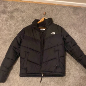Svart dunjacka från The North Face - Säljer en svart dunjacka från The North Face. Jackan har en dragkedja framtill och är långärmad. Perfekt för kyligare väder med sin pufferdesign som håller värmen. Inköp förra året på stadium ej använd.