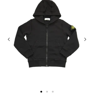 Svart hoodie från Stone Island - Snygg svart hoodie från Stone Island med dragkedja och huva. Tröjan har långa ärmar och en ikonisk logga på ärmen. Perfekt för en stilren och avslappnad look.