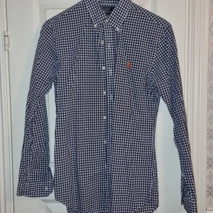 Rutig skjorta från Polo Ralph Lauren - Snygg rutig skjorta från Ralph Lauren i blått och vitt. Skjortan har en klassisk krage och knappar framtill. Den är slim fit och har långa ärmar, perfekt för en stilren look.