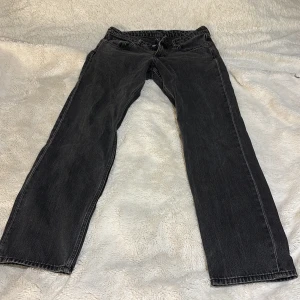 Svarta jeans - Snygga svarta jeans med klassisk low waist design. De har en rak passform och har en ”urtvättad” stil. Byxorna har en knappgylf och bälteshällor. Stl W27/L32. 100cm lång och 40cm bred från ficka till ficka