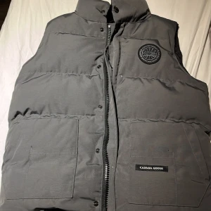 Grå dunväst från Canada Goose - Säljer en stilren grå dunväst från Canada Goose. Jackan har en quiltad design med knappar och flera praktiska fickor. Perfekt för kyliga dagar.