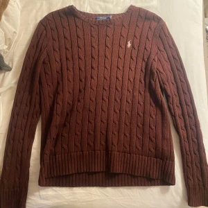 Brun stickad tröja från Ralph Lauren - Säljer en snygg brun stickad tröja från Ralph Lauren med kabelstickat mönster. Tröjan har en klassisk design med rund hals och långa ärmar. Perfekt för kyliga dagar!
