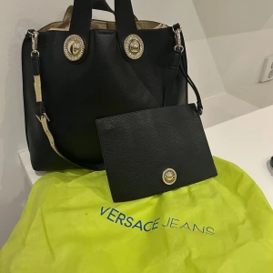 Svart väska från Versace Jeans - Snygg helt ny svart axelväska från Versace Jeans med gulddetaljer. Väskan har en avtagbar axelrem och dekorativa knappar med logotyp. Dustbag medföljer