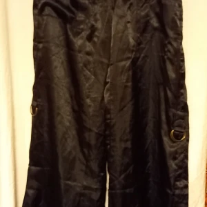 Svarta byxor med ringdetaljer - Snygga svarta byxor i 100% polyester med coola ringdetaljer på sidorna. Byxorna har en loose passform och är perfekta för en avslappnad stil. Plus att man kan dra upp benen med ett band som sitter inne i benen för att fästa i ringen på utsidan. Kommer ifrån E18shoppen