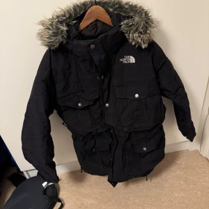 Svart parkajacka från The North Face - Säljer en svart parkajacka från The North Face med pälskantad huva. Jackan har flera praktiska fickor och stängs med både dragkedja och knappar. Perfekt för kalla vinterdagar.