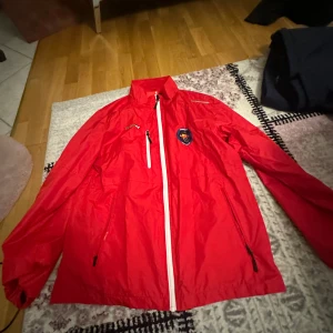 Röd vindjacka från CCM - Säljer en röd vindjacka från CCM med Djurgården Hockey-emblem på bröstet. Jackan har en dragkedja framtill och en hög krage. Perfekt för ishockeyentusiaster! 🏒