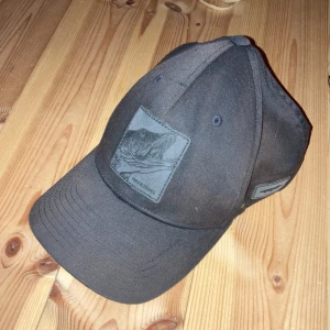 Svart keps från Flexfit - Snygg svart keps från Flexfit med broderad patch framtill. Kepsen har en flexibel passform och är tillverkad i en polyester- och bomullsblandning. Perfekt för en avslappnad stil.(OBS lite solblekt)