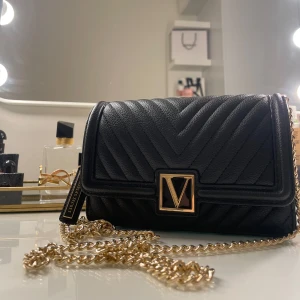 Svart quiltad axelväska från Victoria's Secret - Snygg svart axelväska från Victoria's Secret med quiltad design och guldfärgad kedja. Väskan har en elegant V-logga på framsidan och en praktisk dragkedja inuti för säker förvaring.