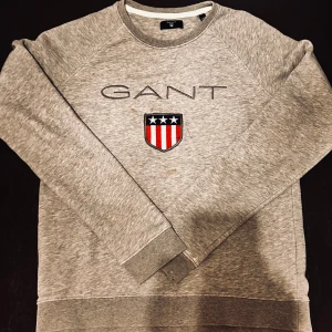 Grå sweatshirt från GANT - Säljer en stilren grå sweatshirt från GANT med broderad logotyp och emblem på bröstet. Tröjan har långa ärmar och rund halsringning, perfekt för en avslappnad look.