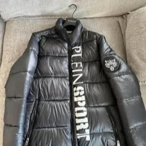 Svart pufferjacka från Plein Sport - Snygg svart pufferjacka från Plein Sport med avtagbara ärmar och stort tryck på framsidan. Jackan har en cool camouflagemönstrad insida och dragkedja framtill. Perfekt för att hålla stilen i kyligare väder. Modell: Dakota  Använd 3 gånger Nypris ca 6000kr