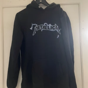Svart Metallica hoodie från Just Hoods - Cool svart hoodie med Metallica-tryck från Just Hoods. Perfekt för fans av bandet! Den har en klassisk passform med långa ärmar och en bekväm huva. Ett måste för rockälskare 🎸