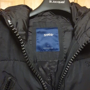 Svart dunjacka från Svea - Snygg svart dunjacka från Svea med pälskantad huva och dragkedja framtill. Jackan har praktiska fickor med lock och ribbade muddar vid ärmsluten. Perfekt för kalla vinterdagar.