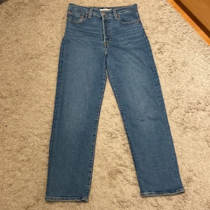 Levi's Ribcage Straight Ankle Jeans - Snygga Levi's Ribcage Straight Ankle jeans i klassisk blå denim. Dessa jeans har en hög midja och rak passform som slutar vid ankeln. Perfekta för en stilren och avslappnad look. Jeansen är använda fåtal gånger och är som nya 