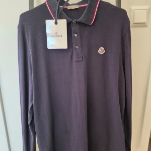 Mörkblå pikétröja från Moncler - Snygg mörkblå pikétröja från Moncler med klassisk krage och knappar. Tröjan har Monclers logotyp broderad på bröstet och röd-vit randdetalj på kragen. Perfekt för en stilren look. Nfc scan, helt ny