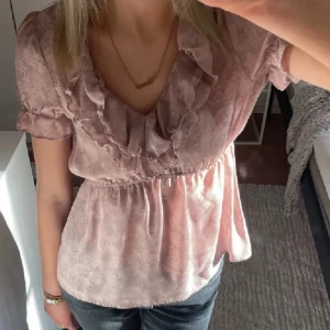 Rosa blus från H&M - Söt rosa blus från H&M med volangdetaljer och korta ärmar. Blusen har en v-ringad design och ett elastiskt band i midjan för en smickrande passform.