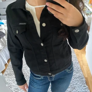 Svart jeansjacka från Calvin Klein - Säljer en snygg svart jeansjacka från Calvin Klein. Den har en cool logga på ryggen och silverfärgade knappar framtill. Perfekt för både vår och höst! Jackan har långa ärmar och en klassisk krage. Passar till alla tillfällen, både casual och lite mer uppklätt.