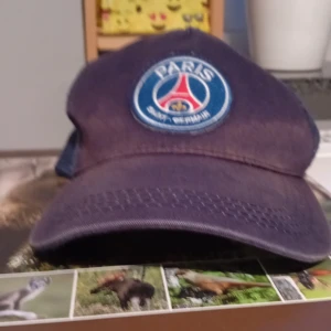 Blå keps med Paris Saint-Germain-logga - Säljer en snygg blå keps med Paris Saint-Germain-logga framtill. Kepsen har en justerbar rem baktill för perfekt passform. Perfekt för sommarens alla äventyr eller för att visa ditt stöd för laget! 🧢