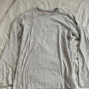 Grå långärmad tröja från Tommy Hilfiger - Säljer en stilren grå långärmad tröja från Tommy Hilfiger. Den är gjord av 100% bomull och har en liten logga på bröstet. Perfekt för både vardag och skola. Tröjan är i bra skick och superbekväm att ha på sig. Passar perfekt till jeans eller chinos!