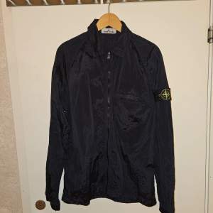 Säljer en exklusiv Stone Island Nylon Metal overshirt  jacka i nyskick. Lätt och bekväm med unik metallisk yta, tvåvägsdragkedja och ikoniskt kompassmärke på ärmen. Perfekt för svala sommarkvällar. Stilrent val för både komfort och design.