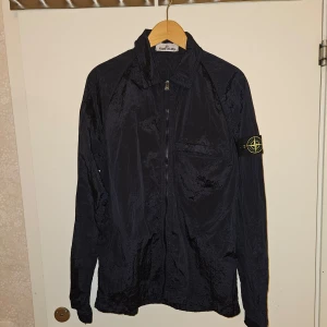 Stone Island Jacka - Säljer en exklusiv Stone Island Nylon Metal overshirt  jacka i nyskick. Lätt och bekväm med unik metallisk yta, tvåvägsdragkedja och ikoniskt kompassmärke på ärmen. Perfekt för svala sommarkvällar. Stilrent val för både komfort och design.