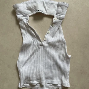 Vit ribbad ärmlös topp - Säljer en stilren vit ribbad topp. Den är ärmlös och har en hög krage med knappar framtill. Perfekt för en somrig look eller att bära under en kavaj för en mer uppklädd stil.