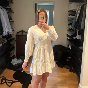 Vit klänning med volangdetaljer - Säljer en superfin vit klänning med volangdetaljer. Den har en v-ringad design och långa ärmar. Klänningen är i nyskick och har bara använts 1 gång.