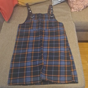 Rutig klänning från Monki - Säljer en superfin rutig klänning från Monki i storlek XS. Klänningen har ett coolt rutmönster i blått, svart och orange. Den är ärmlös med justerbara axelband och en praktisk ficka framtill. Perfekt för höst och vår, och kan stylas med en tröja under för en extra touch!