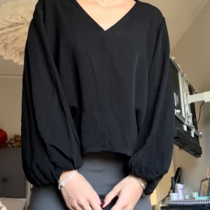 Svart blus från Ivy & Oak - Säljer en elegant svart blus från Ivy & Oak. Den har en v-ringad design och långa, löst sittande ärmar som ger en avslappnad men stilren look. Perfekt för både vardag och fest!