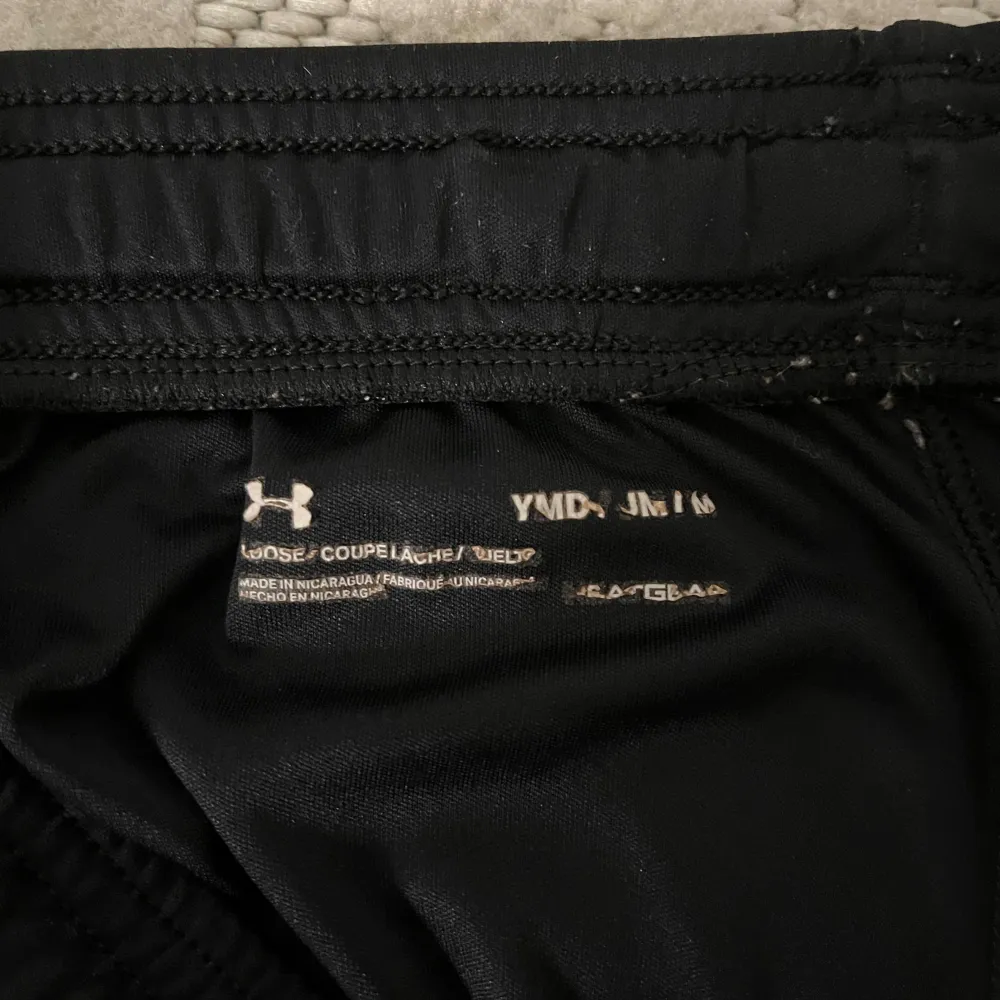 Säljer ett par svarta träningsshorts från Under Armour. De är i bra skick och har en elastisk midja med snörning för bästa passform. Shortsen har en diskret logga på framsidan och 'Under Armour' tryckt längs sidan. Perfekta för träning eller vardagsbruk!. Shorts.