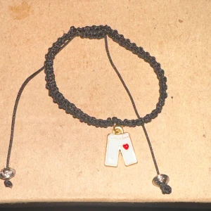 Svart armband med byxberlock - Säljer ett coolt svart armband med en unik berlock i form av vita byxor med ett rött hjärta. Armbandet är flätat och har justerbara knutar med silverfärgade pärlor i ändarna. Perfekt för att ge en rolig touch till din outfit!