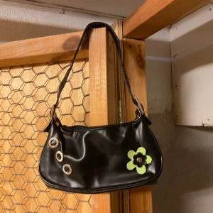 Svart axelväska med blommig detalj - Säljer en snygg svart axelväska i syntetmaterial. Väskan har en cool grön blomma som detalj och tre stora metallringar på framsidan. Den har en dragkedja upptill för säker förvaring. Perfekt för en utekväll eller som en vardagsväska!