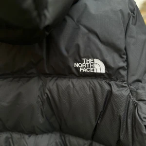 Svart dunjacka från The North Face  - En snygg och varm damjacka i dun från The North Face, storlek M. Jackan är vind- och vattenavvisande och har en praktisk huva för extra skydd mot kyla. Resor i ärmslut, dragkedja framtill. Perfekt för kalla vinterdagar! Jackan är ny, utan taggar dock